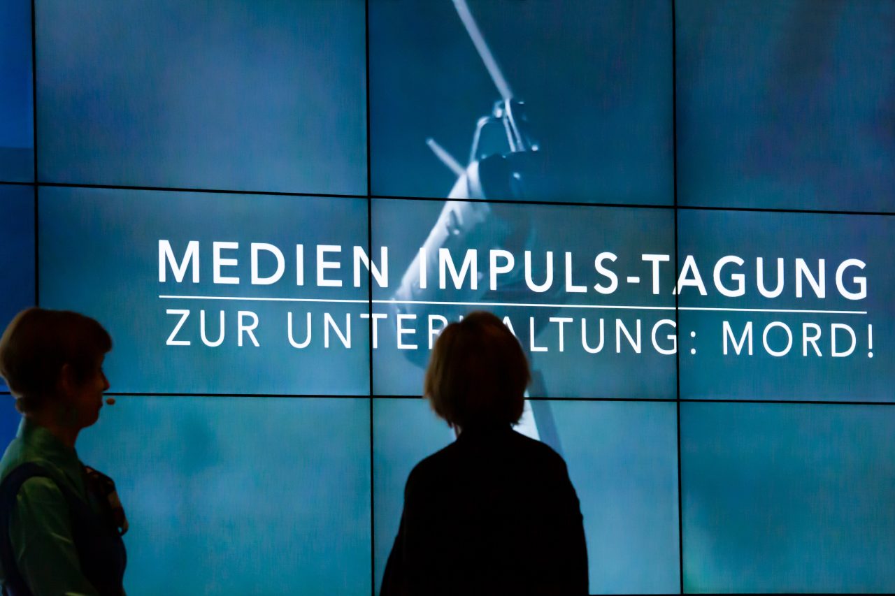 Silhouetten zweier Personen vor einer großen Medienwand. Die Medienwand zeigt ein dunkles Bild und den Titel der Veranstaltung.